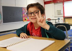 Alexzandr Patti, a young child who has short brown hair and is wearing oversized eyeglasses, a red t-shirt and green hoodie, looks at and smiles for the camera while making a peace sign with their fingers. Zandr is seated at a desk in a classroom with a book open on the table they are seated at.
