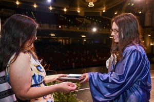 An individual hands a student dressed in blue graduation regalia a plaque on stage.