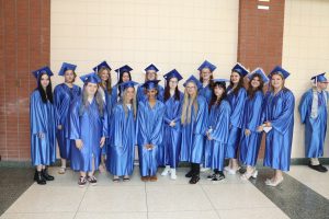 A group photo of 15 students in blue graduation regalia huddled, smiling.