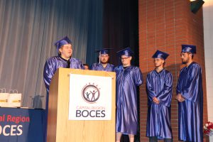 A student in blue graduation regalia speaks at a podium, while four students also in blue graduation regalia stand next to them.