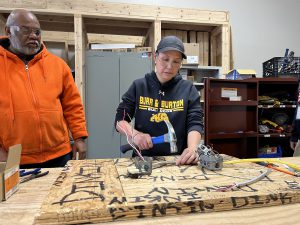 Two adult students in a workshop, one standing in an orange hoodie as the other in a black hoodie and a hat uses a hammer.