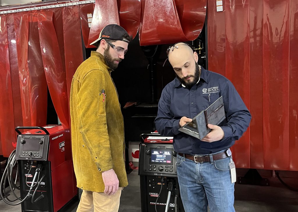 An adult welding student in a yellow safety coat looks on as an instructor demonstrates with a tool.