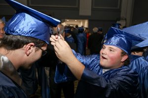 Two students in blue graduation regalia, one student fixes the tassel of the other student.