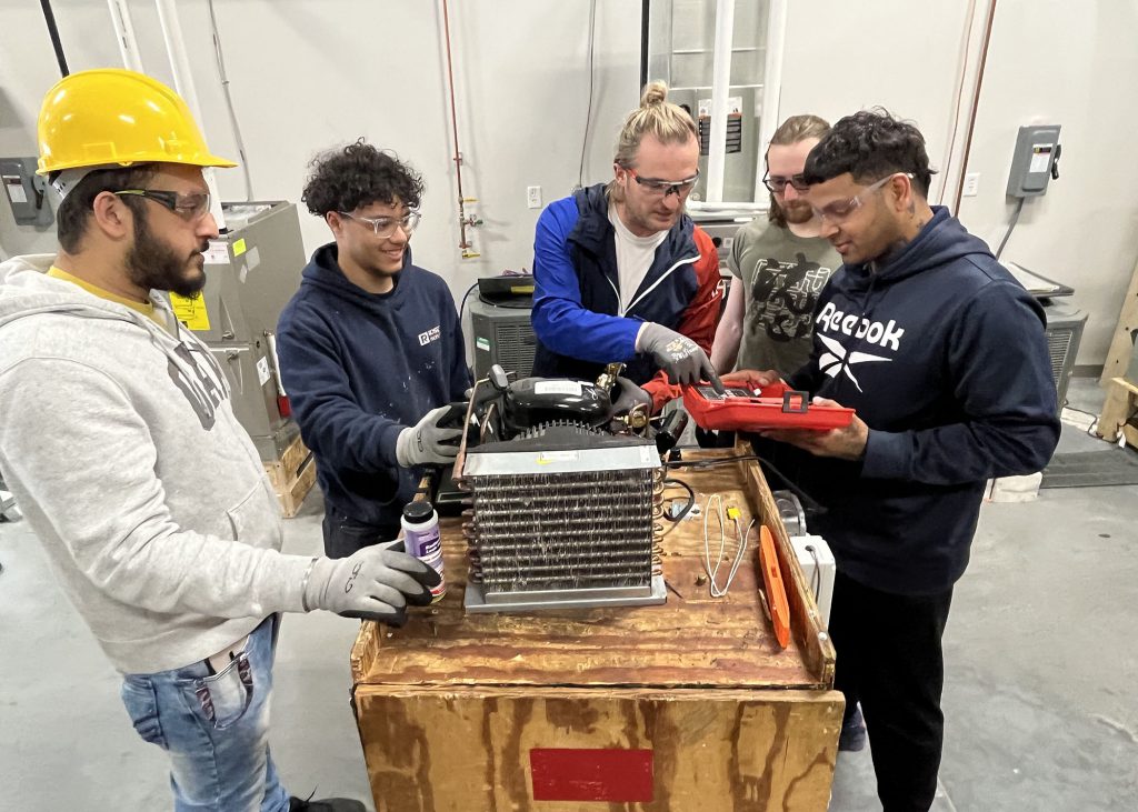 Five adult HVAC-R students stand over refrigeration equipment. Two students are working with tools while the other 3 observe.