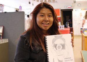 High schooler Joselyn Diaz Garcia, who has long reddish brown hair, is wearing a black denim jacket and is displaying a copy of the Creative Collective writing anthology with their artwork on its front, looks at and smiles for the camera. 