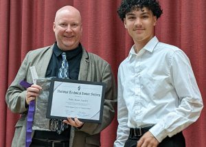 Culinary student Pedro Sanchez, who has dark, curly hair and is wearing a white button down shirt and black pants, stands at right next to a Career and Technical Education teacher who is holding a National Honor Society certificate. Both are looking at and smiling for the camera. 