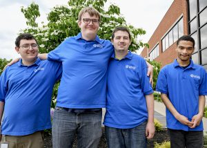 Four young adult students who are graduating from the Work Based Learning program stand in a row with arms around each other's shoulders. They are outside the entrance to Capital Region BOCES. All are wearing bright blue polo shirts and are looking at and smiling for the camera.