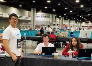 A person in a t-shirt stands in front of a table where two other students are sitting.