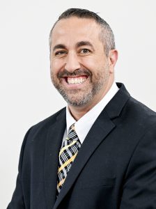 Maywood Academy Principal Gabriel Barbato, who has close cut black and grey hair, a trim beard and mustache and is wearing a white collared shirt, black and yellow plaid tie and black suit coat, looks at and smiles for the camera.