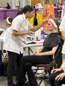 A cosmetology student stands applying makeup on another seated in a chair.