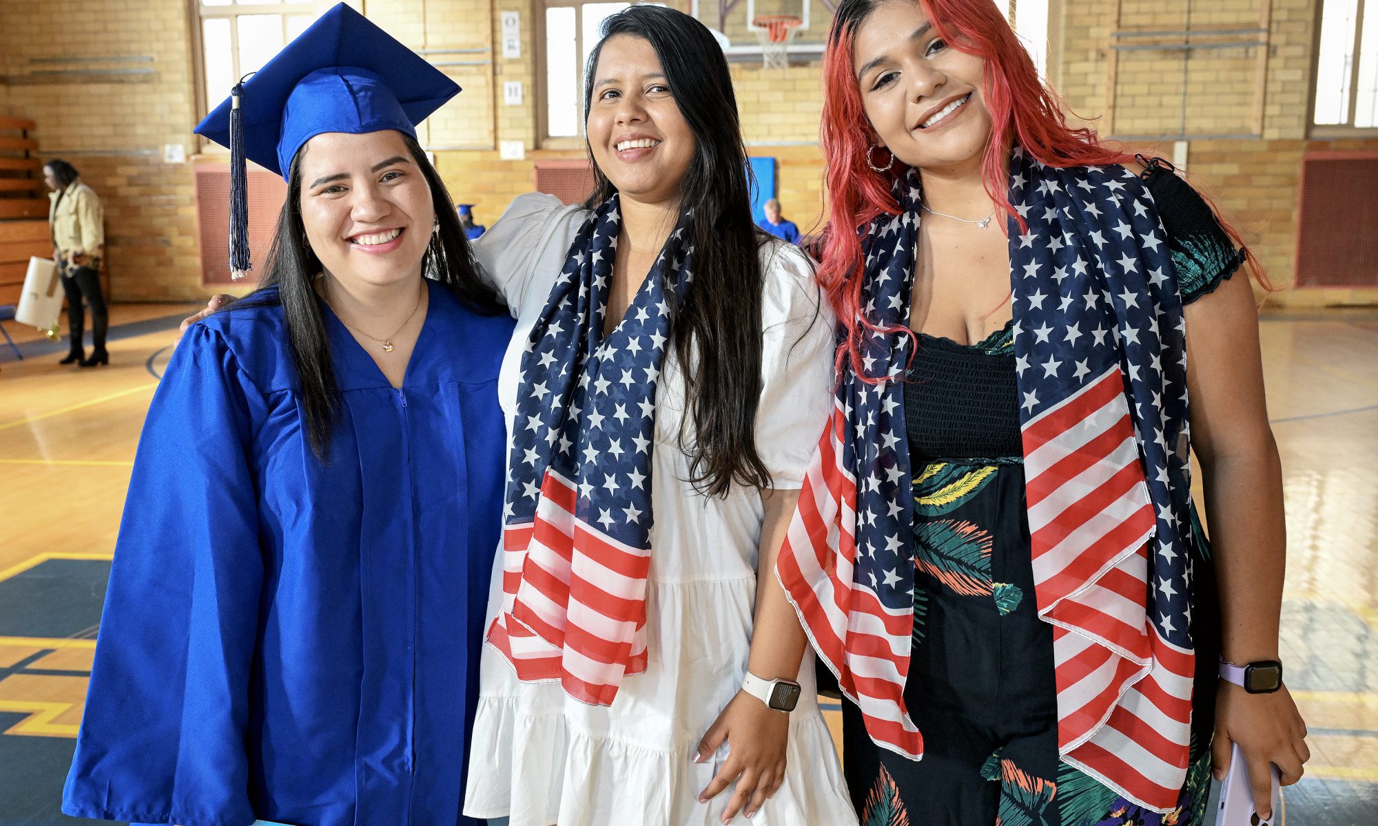 Three woman stand together to pose for a picture. One on the far right is dressed in a graduates cap and gown. The other two each ware scarves depicting the American flag.