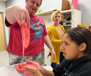 A teacher looks on as one student slowly drops a gooey pink substance into the cupped hands of another student.