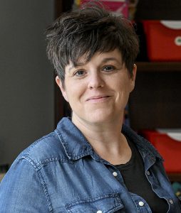 Airline Drive Academy Assistant Principal Amanda McNeal, who has cropped black blond hair and is wearing a black shirt and blue button-up shirt, looks at and smiles at the camera while standing in front of a bookshelf.