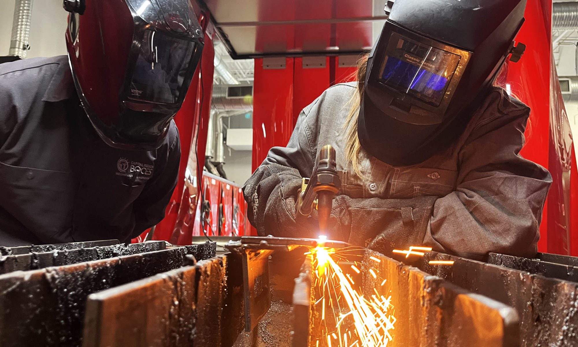 The faces of a student and teacher are hidden behind welding helmets as sparks fly between them as they work.