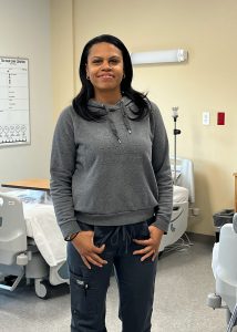 A student wearing a grey sweatshirt and blue jeans stands in front of a patient's bed at the campus' mock hospital room at Capital Region BOCES.