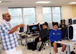 A teacher stands in front of three students seated in front of classroom computers in a game design class.