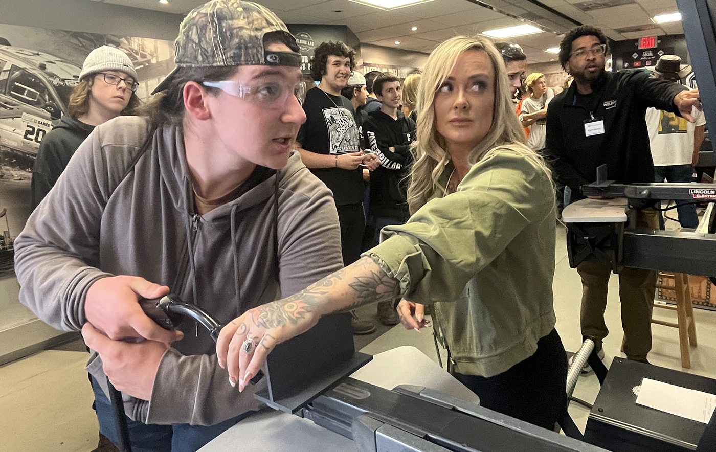 Two students working together on a piece of machinery as classmates look on in the background in Welding and Metal Fabrication class.