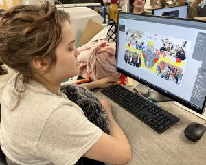 A female student in a tan t-shirt sits down at her desk while looking over the details of a design on a computer screen.