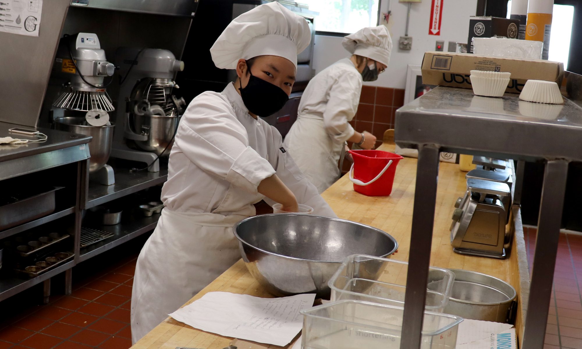 A chef wearing a white outfit, apron and hat, works behind a counter as she prepares food.