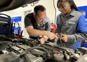 A student technician and her instructor lean over a car engine together.
