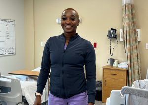 A student wearing a navy blue shirt and purple pants stands in front of an empty patient's bed in the campus' mock hospital room at Capital Region BOCES.