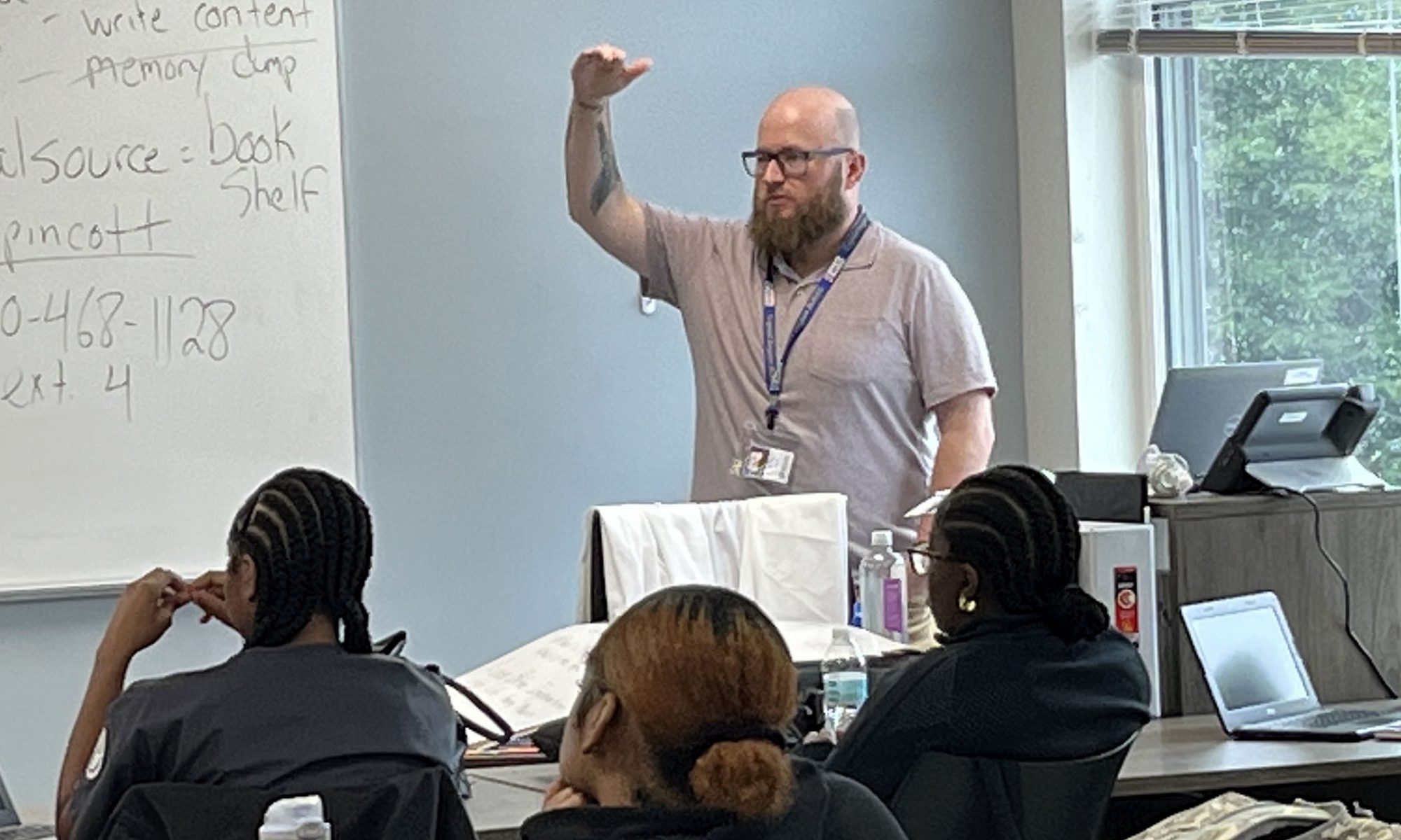 Teacher wearing a grey polo shirt stands with his right hand up high to demonstrating the height of something as he speaks before a classroom of students.