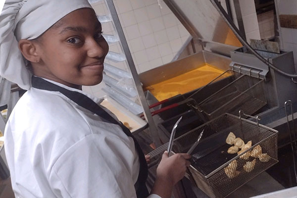 A female student in traditional chef garb stands in front of a deep fryer with a basket of food hovering over the oil.