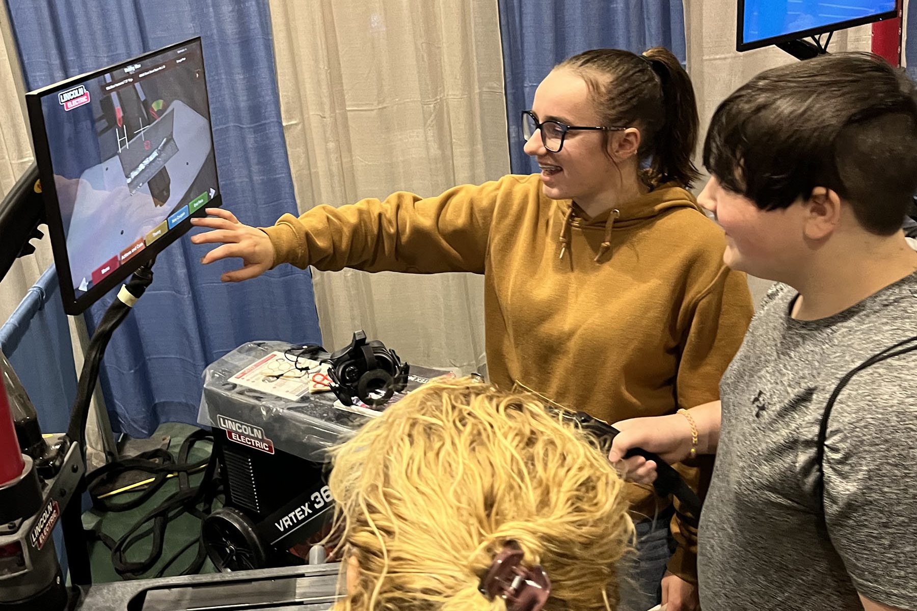 Two students look over a computer screen, while one handles the controls on an apparent computer program that simulates the operation of heavy equipment.