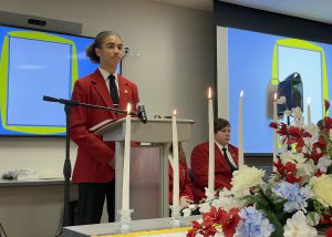 A male student in a red coat stands behind a podium, calling a special meeting to order.