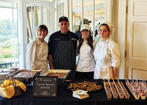 Four people, two chefs and two students, stand before a table displaying various prepared foods.