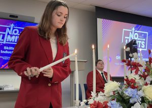 A female student in a red jacket stands behind a candle, lighting the wick in a ceremony.
