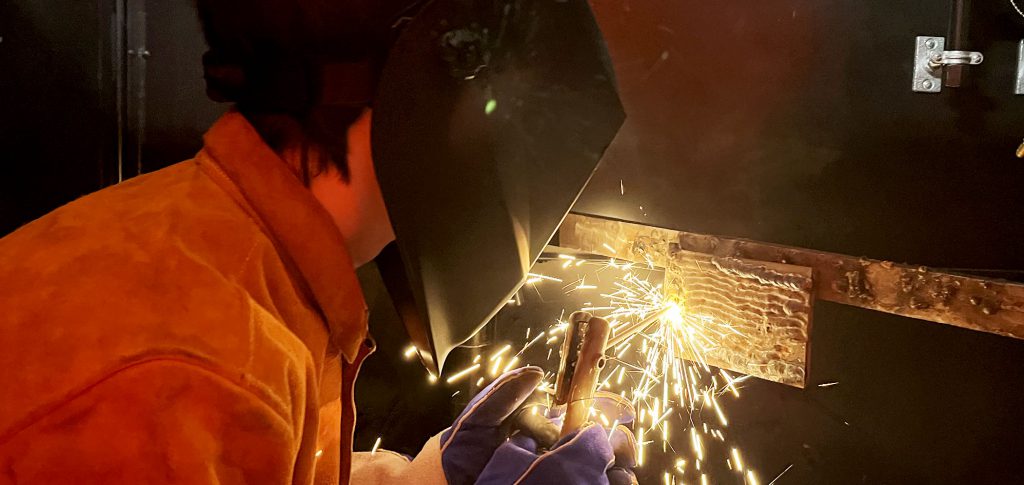 A student in a welding helmet works with sparks flying from her tools.