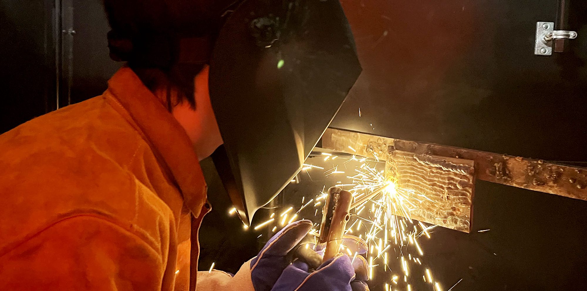 A student in a welding helmet works with sparks flying from her tools.