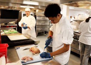 A male student wearing traditional chef garb, sans hat, works on breaking down chicken to prepare it for cooking.