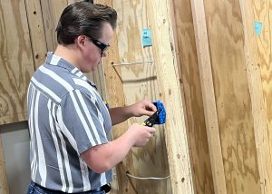 A student in safety goggles works on installing an electrical outlet on a wooden supportive strut.