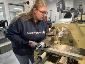 A female student in a long sleeved blue shirt, her long blond hair tucked behind her, is working on a piece of woodcutting machinery.