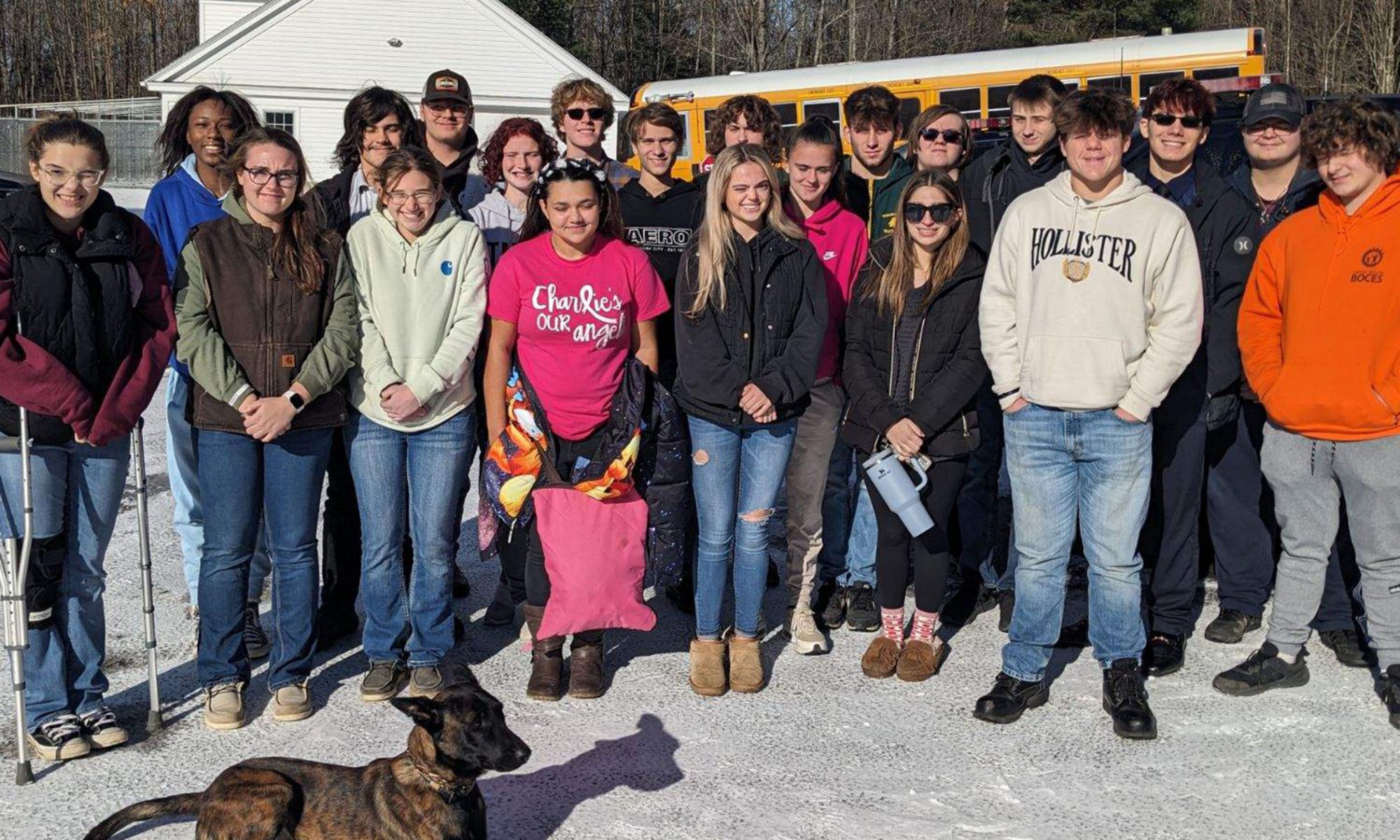 A group of students stand in the background behind an attentive police dog laying down on the ground.