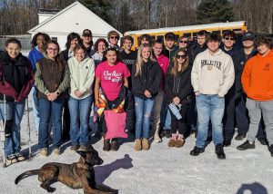 A group of students stand in the background behind an attentive police dog laying down on the ground.