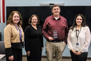 Capital Region BOCES administrators Lauren Gemmill, Monica Lester and Elizabeth Wood stand in a row with Public Information Specialist Zach Ribert, second from right. Ribert is holding a clear glass service award. All are looking at and smiling for the camera.