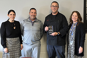 Capital Region BOCES administrators Lauren Gemmill, Jeff Palmer and Elizabeth Wood stand in a row with teacher Chris Snye, second from right. Snye is holding a clear glass service award. All are looking at and smiling for the camera.