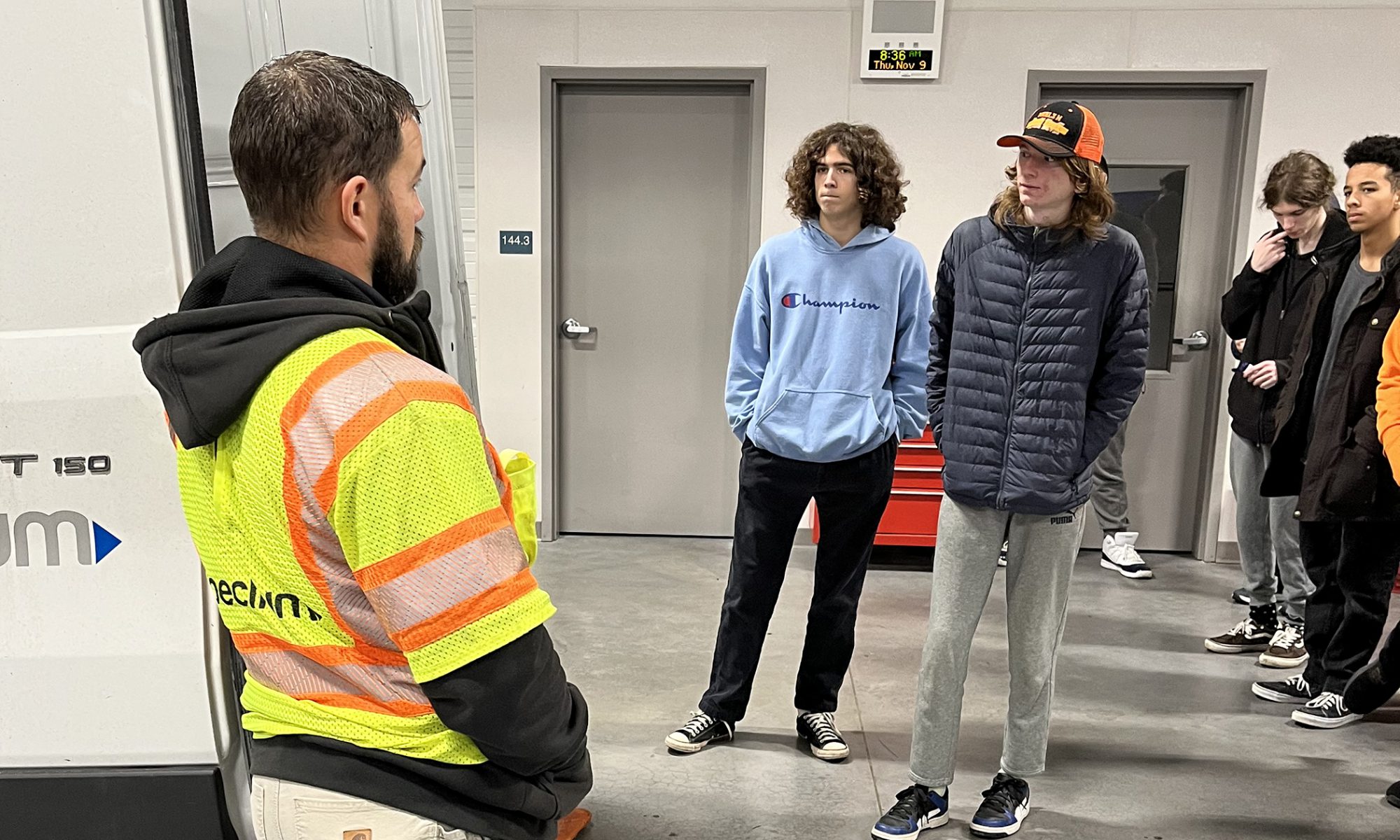 A group of students casually stand as they peer inside a service van while someone in a bright yellow safety vest speaks.