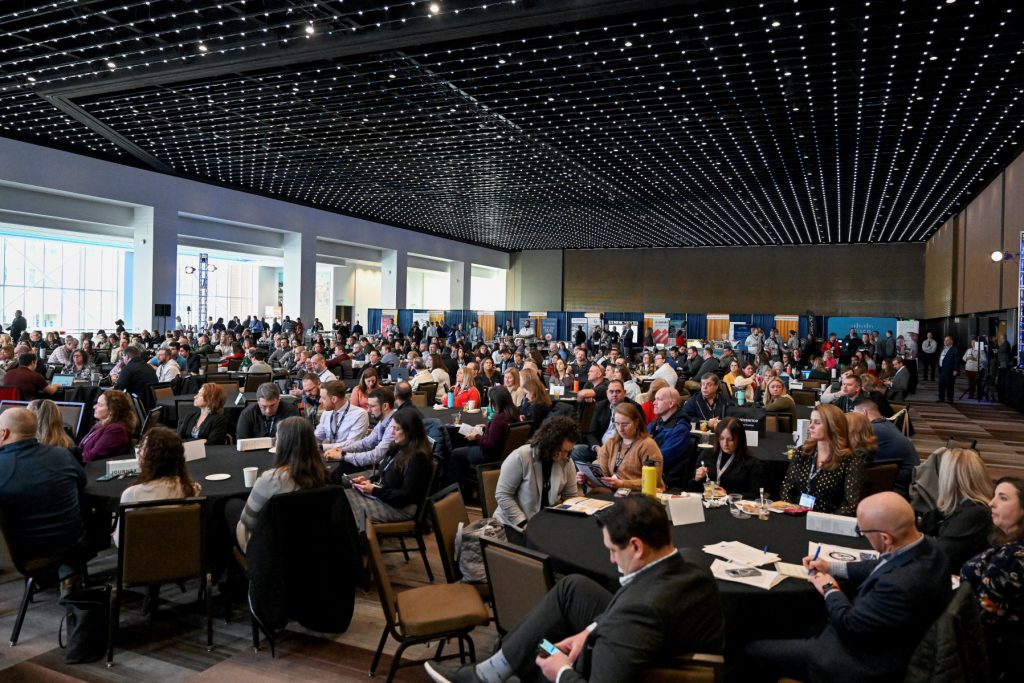A crowd of more than 500 people seated in a large room listening to a presentation off camera.