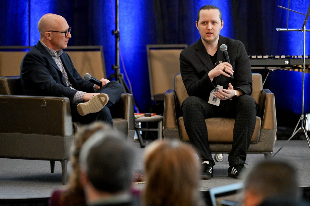 Two gentleman seated on stage, one listening intently while the other speaks into the microphone.