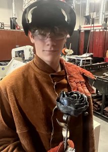 A student in a welding helmet atop of his head holds up a metal rose.
