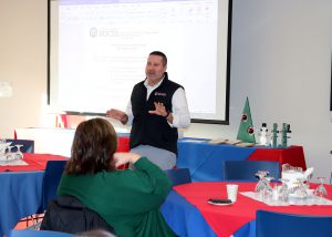 A man in a sweater vest speaks in front of an audience.