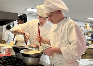 Two culinary students in traditional chef attire work on stirring the contents of a pie filling.