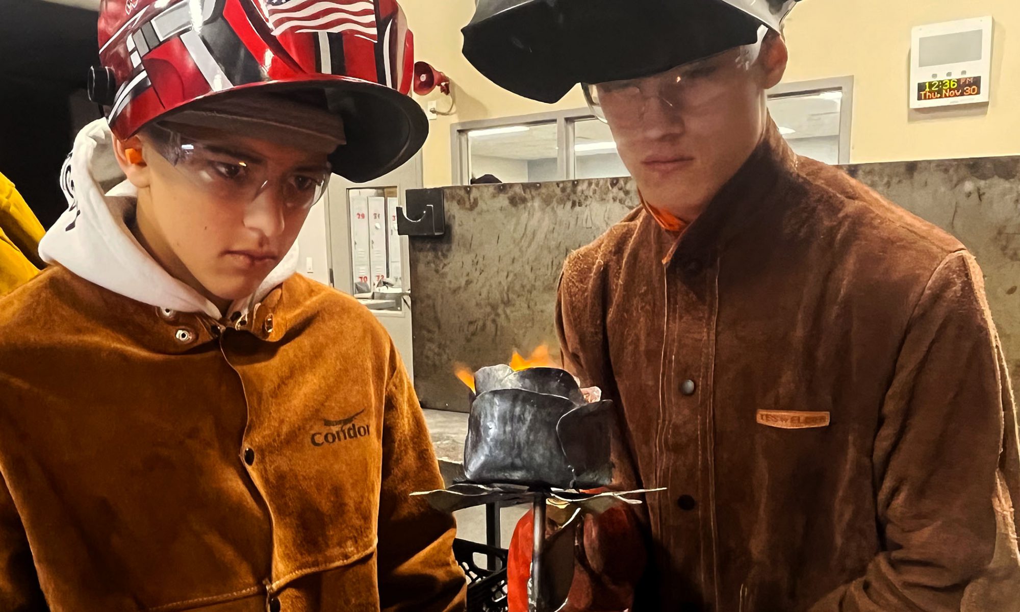 Two students in welding helmets hold a freshly fabricated metal rose that still carries a flame.