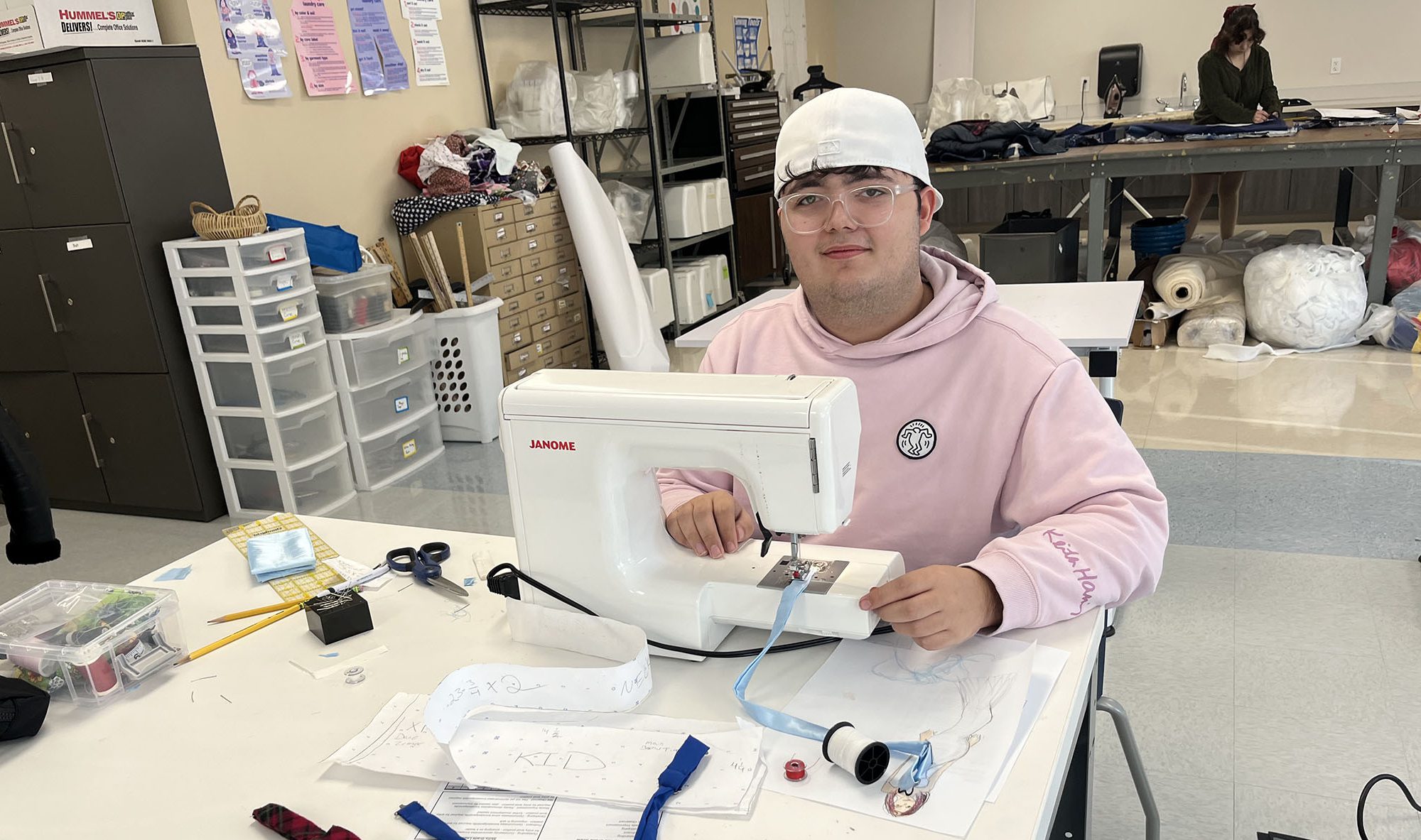 A young man wearing a white, backwards baseball cap and a pink hoodie sits at a sewing machine with a display of various bow ties arranged in front of him.