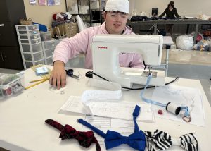 A young man wearing a white, backwards baseball cap and a pink hoodie sits at a sewing machine with a display of various bow ties arranged in front of him.
