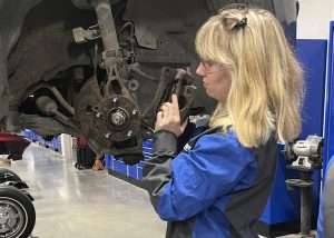 A student has her eyes on the wheel assembly of a car that has been hoisted on a lift inside the garage.